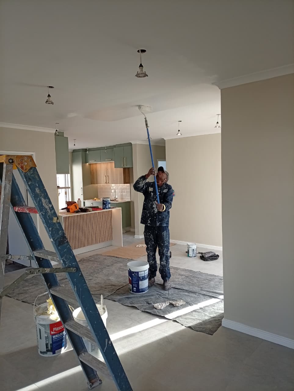 Painting the ceiling of an open-plan living area with an extension roller, sage-green kitchen cabinetry in the background