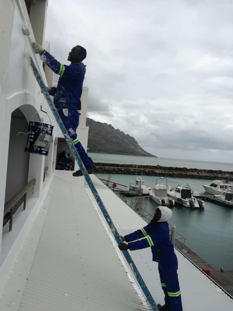 Two workers painting the exterior wall of a harbourside building from a ladder, boats and mountains behind them